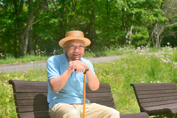 An elderly man in a khaki jacket and hat, holding a walking stick and sits relaxed under the tree’s shade in a public park.