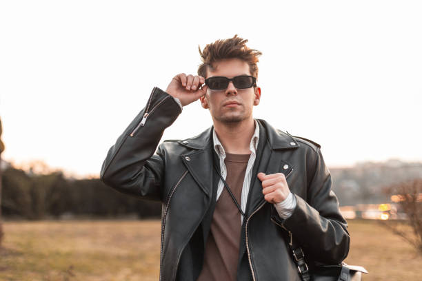 istock Fashionable young hipster man in vintage leather oversized black jacket with backpack straightens sunglasses outdoors. American guy in trendy casual clothes posing in nature. Retro fashion. 2154712474