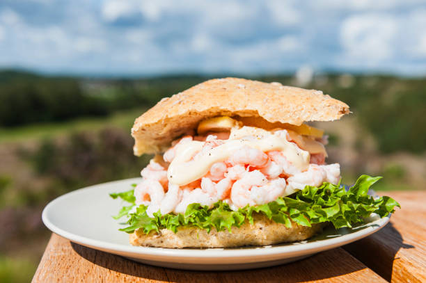 A Swedish shrimp sandwich rests on a white plate placed on a wooden table. The sandwich is neatly arranged, ready to be eaten. The texture of the bread and fillings is visible against the clean white plate.