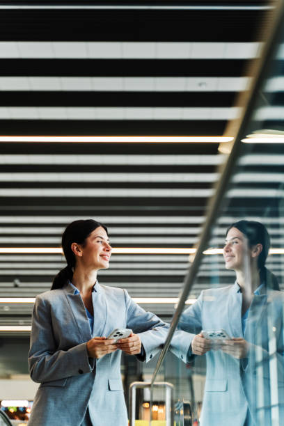 istock Tourist with smartphone, documents and backpack in the airport lounge is looking through the panoramic window. Smiling caucasian woman waiting for flight to start vacation in 2024. 2154733982