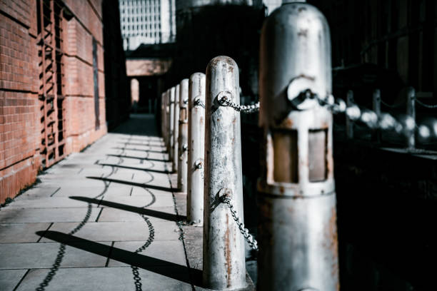 An Urban pathway with metal posts and chains, long shadows. Brick buildings create an industrial, gritty atmosphere.