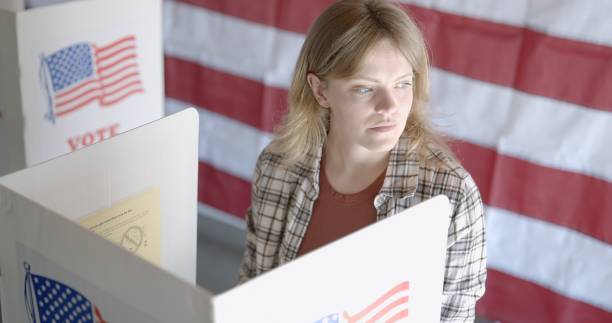 istock Medium shot, young woman thinking at voting booths, US flag behind 2154771521