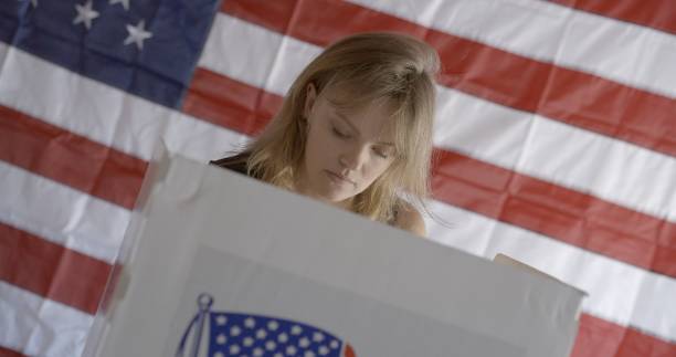 istock Angled frontal view, young woman at voting booth, US flag behind 2154771532