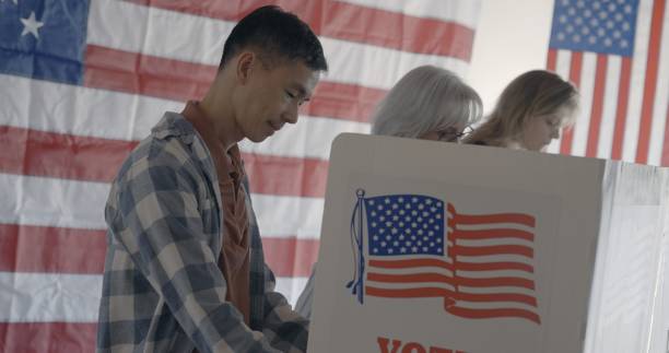 istock Asian man votes at election booth with other voters and US flag behind 2154771555
