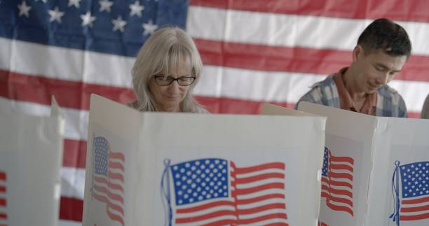 istock Frontal view voting booths with focus on senior woman, US flag behind 2154771637