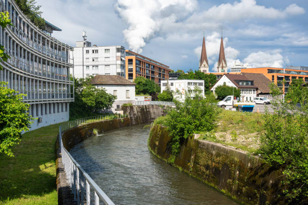 Urban scene in Olten with glass building by river Dunnern, traditional and modern houses, church spires, lush greenery, serene atmosphere.