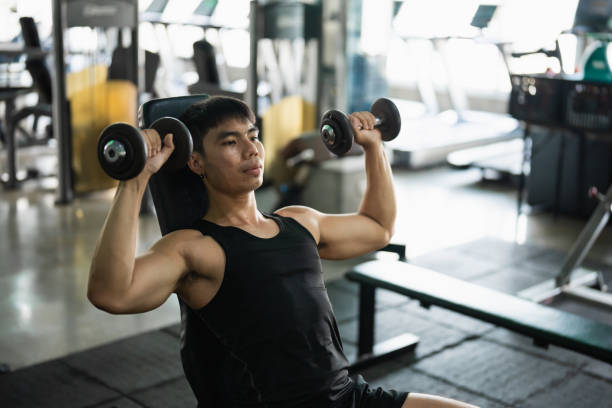 A focused man in a black tank top performs a seated dumbbell shoulder press in a modern gym. His muscular build and concentrated expression highlight his dedication to strength training.