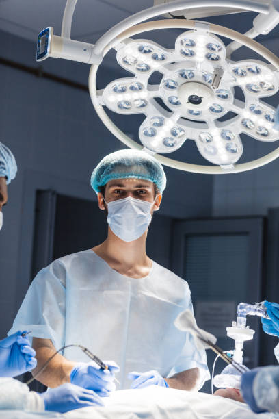 Portrait of young doctor wearing medical protective mask and scrub cap. Practicing surgeon in sterile surgical wear, bright operation room background.