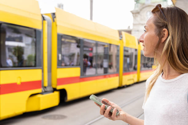 A woman with sunglasses on her head and a backpack checks her smartphone while waiting at a tram station, with a yellow tram in the background.