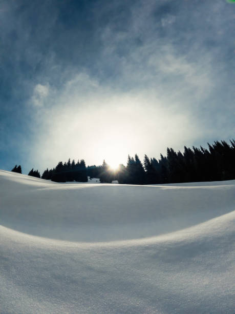 A wintry scene with snowcovered mountain slopes, trees in the background, and the sun peeking through the clouds, creating a picturesque natural landscape.
