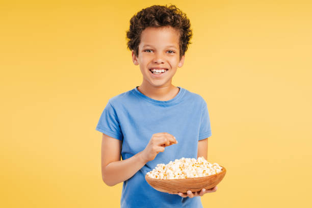 Smiling handsome African American boy watching movie, eating popcorn, snacking looking at camera standing isolated on yellow background. Concept of entertainment