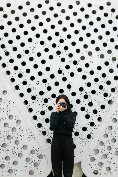 Young woman wearing a black clothes taking a photograph with an old film camera, standing in front of a white wall with black polka dots, creating an artistic backdrop.
