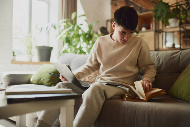 istock Involved in studying. Concentrated young guy in beige outfit sitting on sofa at home, looking in book, using laptop, doing homework tasks. 2154838145