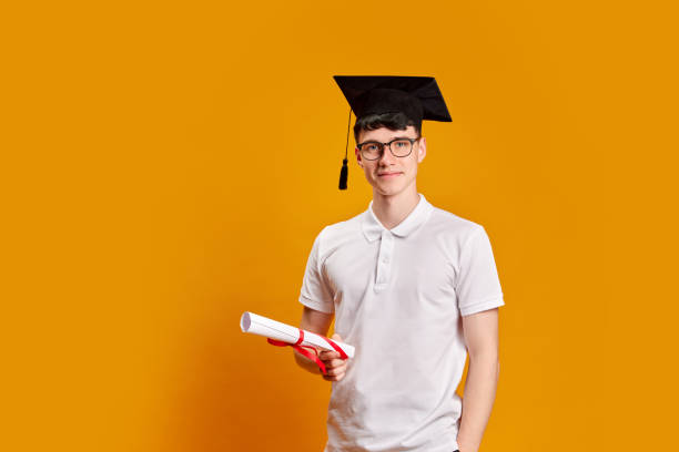 Young man, student in graduation cap, complete education with graduation regalia and diploma against bright yellow background. Concept of education, studying, homework, youth, lifestyle