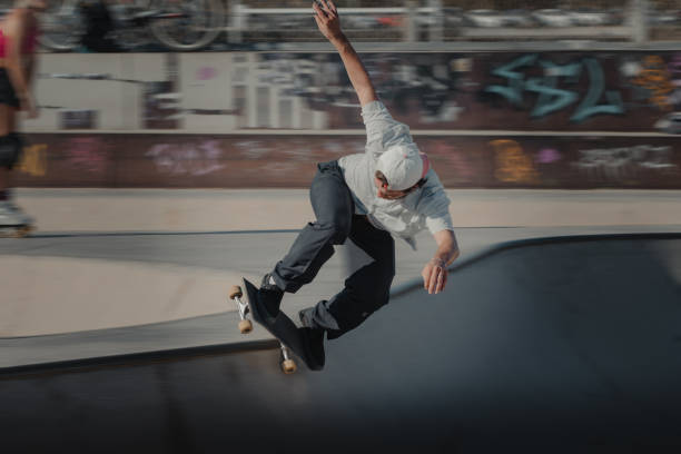 A person skateboarding in a skatepark. The skater, wearing a white cap, white t-shirt, and dark pants, is captured mid-air while performing a trick on the curved surface of the ramp. The background is blurred due to motion, emphasizing the dynamic speed of the scene. Graffiti is visible on a wall behind the skater, adding to the urban atmosphere of the skatepark. Another person is seen in the background on the left side of the image, but is not in focus. This image is compelling as it captures a dynamic moment in action sports and conveys a sense of freedom and skill associated with skateboarding culture.
