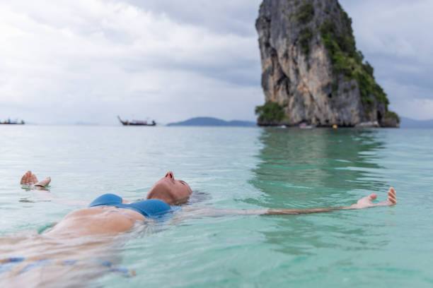 A woman in a blue bikini floats peacefully in clear turquoise waters with a towering rocky island in the background.