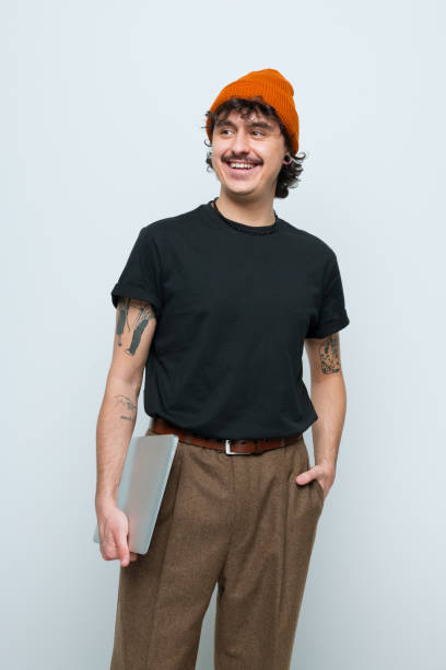 Portrait of friendly young man Confident gen Z young man wearing black t-shirt and orange beanie holding laptop, looking away and smiling. Studio shot, white background. young men stock pictures, royalty-free photos & images
