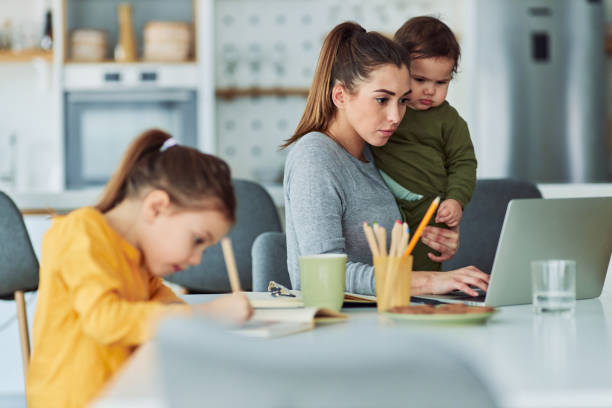 A focused single mother using a laptop and working from home while taking care of her kids A focused single mother working from home on her laptop and taking care of her kids while sitting at a table together. man work from home baby stock pictures, royalty-free photos & images