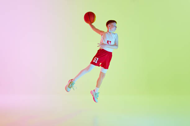 istock Dynamic full-length image of bot in uniform, basketball player practicing, jumping with ball held in one hand against gradient background in neon light 2154879398