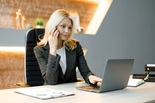 Focused smart surprised successful influential caucasian businesswoman, freelancer or top manager sitting at table in an office using laptop, talking by cellphone, browsing internet or e-documents enfocada inteligente sorprendida exitosa empresaria caucásica influyente, autónoma o alta gerencia sentada a la mesa en una oficina usando una computadora portátil, hablando por teléfono celular, navegando por internet o documentos electrónicos