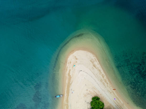 Aerial shot of a sandy peninsula surrounded by clear blue waters, featuring a moored boat and minimal human presence.