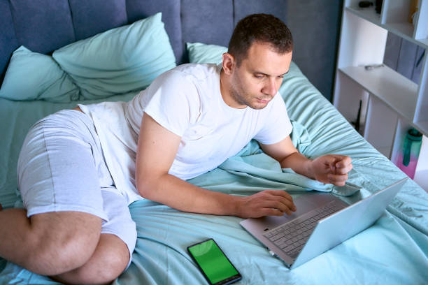 istock a young man is working on a laptop and a smartphone in bed, a cat is lying next to him 2154907014