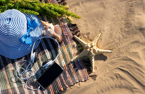 On a bright summer day, a striped plaid with a blue striped summer hat, a large shell, a starfish and headphones with a phone lies on the sand.