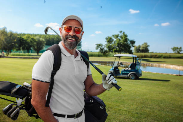 Happy mature golfer standing on the field and smiling to the camera Happy Caucasian mature golfer standing on the field, carrying his golf club and bag, and smiling to the camera hot handsome man stock pictures, royalty-free photos & images