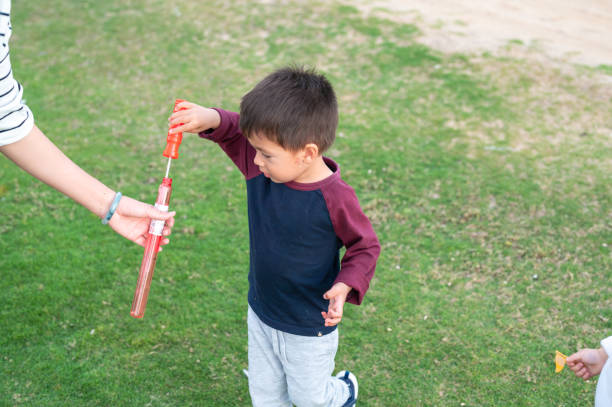 A 3 year old boy is engrossed in playing with a bubble wand in a lush, green park. He holds the wand carefully as he prepares to blow bubbles, capturing a moment of simple joy and childhood wonder.