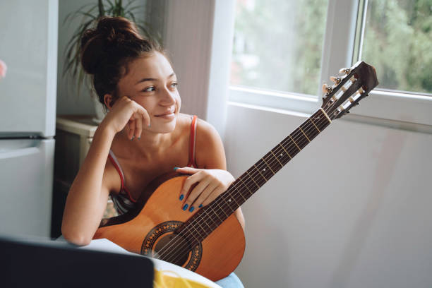 A thoughtful young girl casually holds a guitar, looking away, with natural lighting