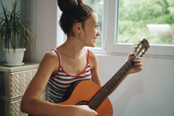 Smiling woman sits in a window and plays the guitar while staying at home. Self isolation concept.