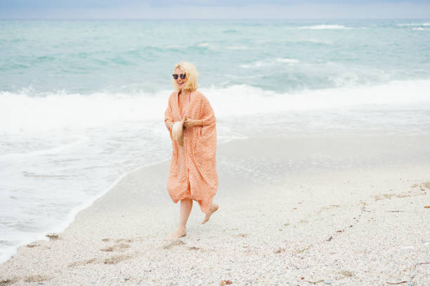 A joyful woman enjoying a relaxing day on a sandy beach by the ocean. She is wearing a flowing dress and sunglasses.