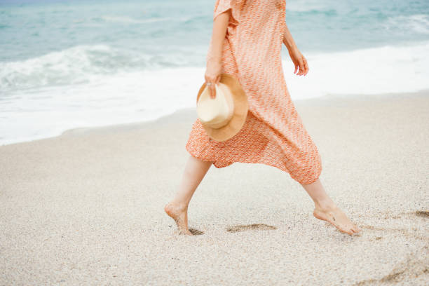 A relaxing scene of a woman walking barefoot on a sandy beach while holding a hat, enjoying a peaceful moment.