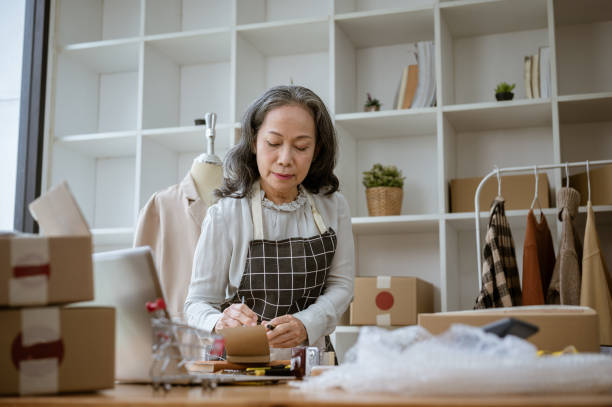 istock A mature Asian female tailor is taking notes on a notepad, measuring and adjusting the pattern. 2154968226
