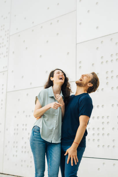 Couple laughing together while standing against a modern white wall with circular patterns. They are clearly enjoying each other's company and sharing a joyful moment.