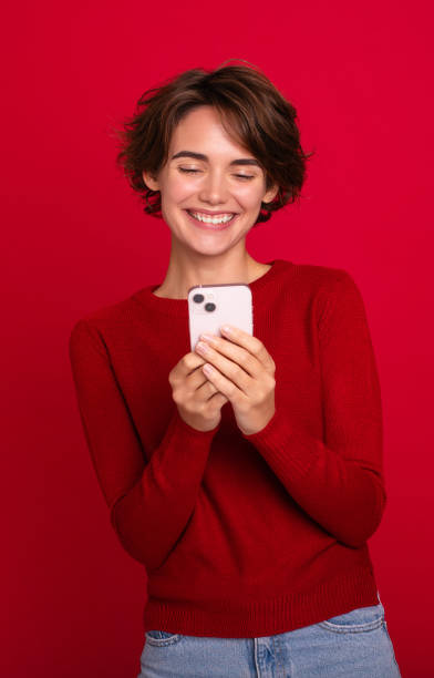 istock Vertical photo of a smiling beautiful young modern woman who using a smartphone isolated on a red bright wall. Shopping, chatting, surfing the internet, betting, social media, win in games 2154978310