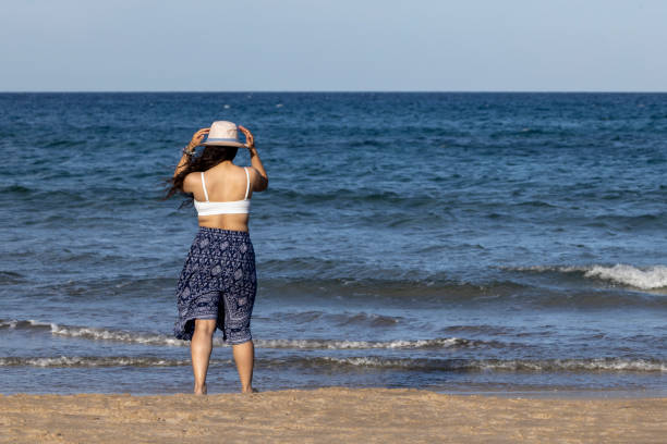 Girl from behind on the beach, touching her hat. Wearing a dress and white bikini. In summertime a sunny day.