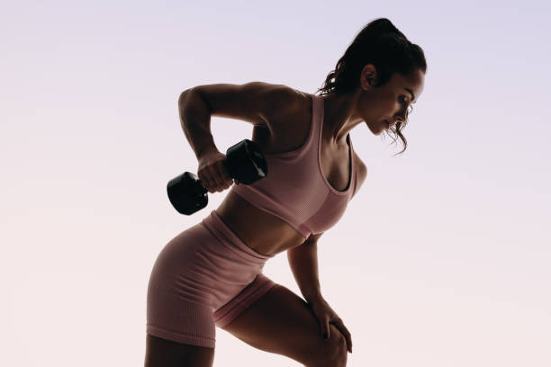 Female fitness: Toned woman listing a dumbbell in a studio workout Female athlete in fitness clothing performs a weightlifting routine in a studio. This woman showcases strength and muscle building, with a lean, toned physique reflecting a healthy lifestyle. dumbbell silhouette stock pictures, royalty-free photos & images