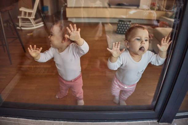Adorable toddler wearing a white outfit is intrigued by their reflection in a sunlit window. Perfect depiction of childhood curiosity and discovery.