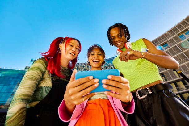 Three Gen Z friends using a smartphone together. Low angle shot with modern buildings in the background. Social media and friendship concept. Three young friends using a smartphone together. Low angle shot with modern buildings in the background. Social media and friendship concept. fish eye lens stock pictures, royalty-free photos & images