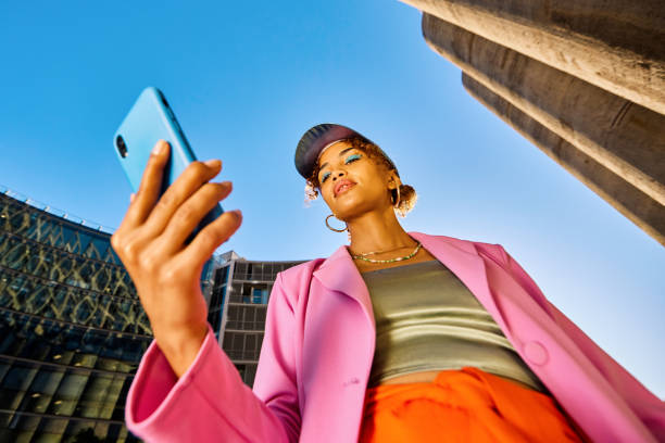 kuvapankkikuvat ja rojaltivapaat kuvat aiheesta young african american woman in colorful clothing using a smartphone. low angle shot with concrete silos in the background. urban fashion and technology concept. - pitää sylissä