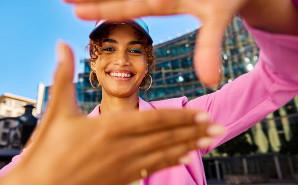 Gen Z woman framing her face with her hands and smiling. Close-up shot with modern building in the background. Positive emotions and urban lifestyle concept. Young woman framing her face with her hands and smiling. Close-up shot with modern building in the background. Positive emotions and urban lifestyle concept. african american architecture stock pictures, royalty-free photos & images