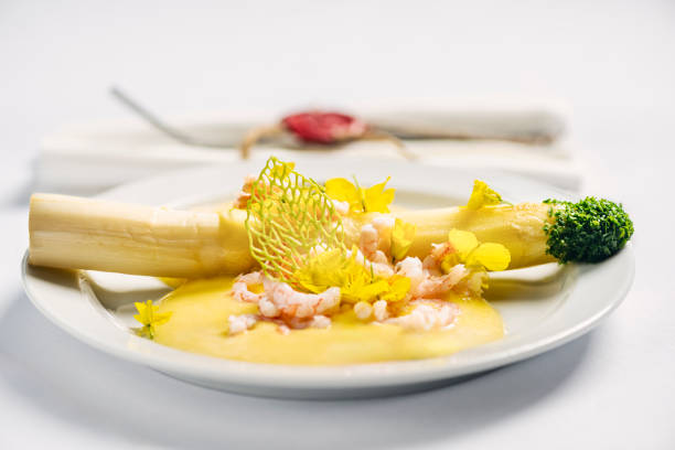Close-up of a dish of white asparagus with a prawn and hollandaise sauce, served with parsley-infused wafer tuile, and organic canola flowers. The tip of the asparagus stalk has been covered with dried dehydrated parsley leaves.