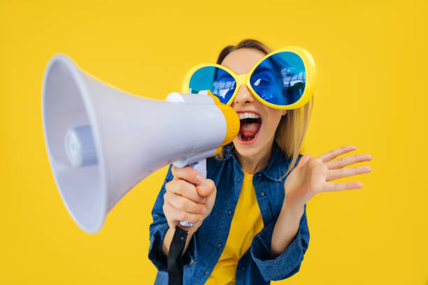 istock Active woman in comic glasses holding loudspeaker and making advertisement while laughing. Energetic young woman entertaining people while telling funny jokes in megaphone over yellow background. 2155028576
