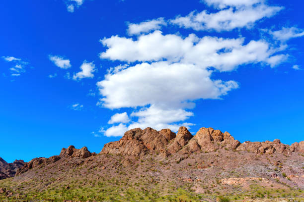 Rugged mountains, composed of reddish-brown rocks and sparsely dotted with vegetation, rise majestically from a flat terrain under a blue sky with sparse clouds in Nevada.
