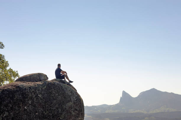 istock A man seated on a cliffside rock, overlooking mountain peaks and a verdant valley. 2155038781