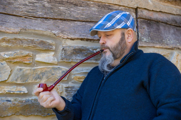 Close up of a man wearing a blue plaid cap smoking a red pipe while leaning against a rock wall.