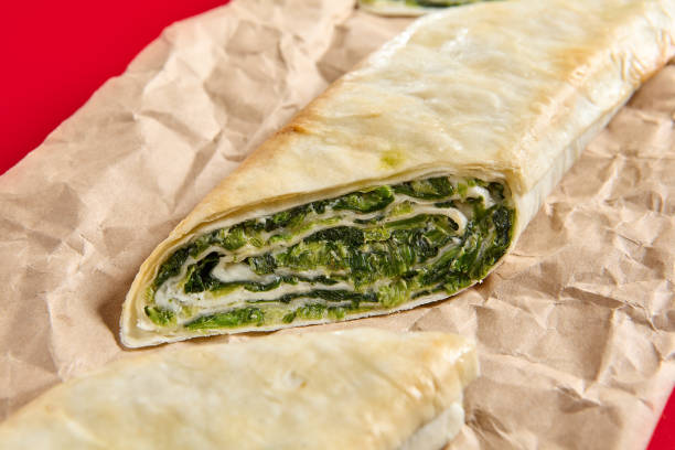 A detailed close-up shows a savory spinach and cheese filled pastry roll with a crispy crust, presented on parchment paper.