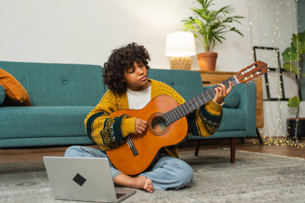 African American woman learning guitar watching tutorial on laptop at home. Black girl playing guitar singing song learning online music lessons, Artistic woman playing acoustic guitar learning chords