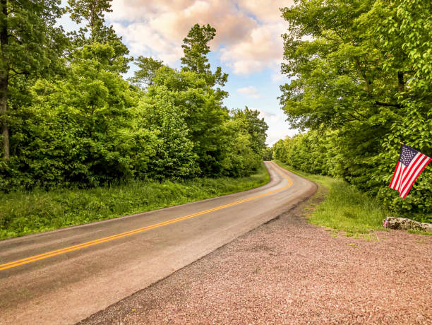 An American flag is waving in the breeze along the road outside Grantsville Maryland.  The country road is curving and is surrounded by lush trees on both sides.  There is a gravel driveway connecting to State Route 495 in Garret County;